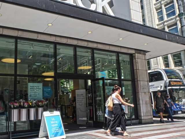 Shoppers leaving Nordstrom Rack. Pedestrians coming around a corner. Bus and buildings.