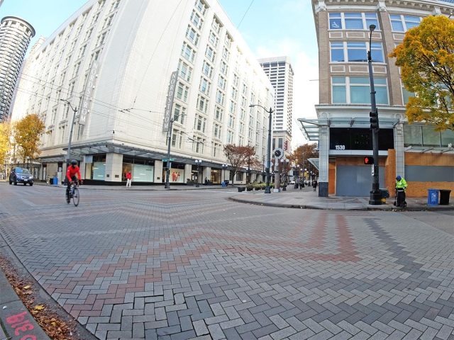 A quiet, mostly empty street in downtown Seattle: one biker, a MID ambassador, one car. Brick road with greys and reds. Tall white building and blue sky.