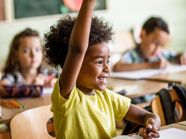 Young black student sitting at a desk, raising his hand and smiling