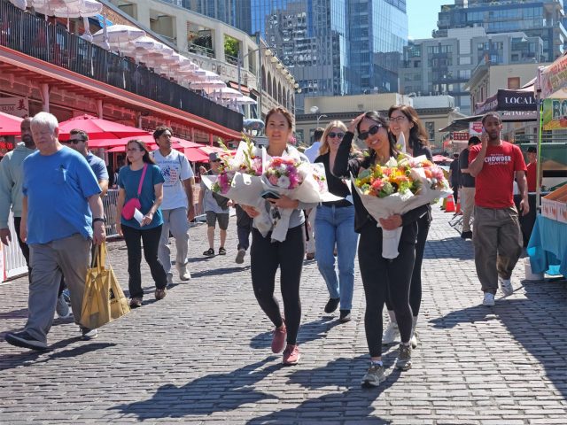 Pike Place Market visitors walk on the cobblestone, smiling and carrying flowers.